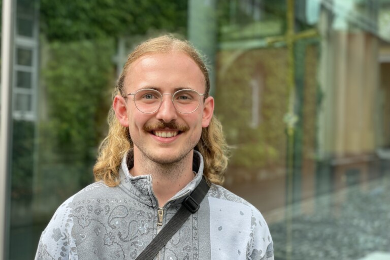 Ein junger Mann mit langem blondem Haar und Brille lächelt in die Kamera, im Hintergrund eine verschwommene städtische Umgebung.