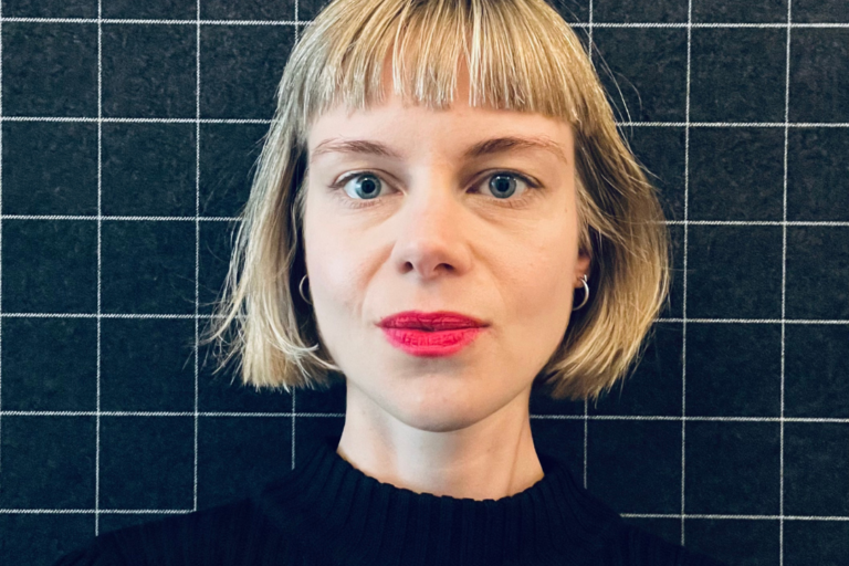 Porträt einer Frau mit blondem Bob-Haarschnitt und rotem Lippenstift, vor einem karierten Hintergrund.
